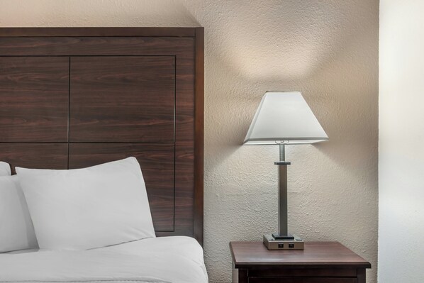 A bed with white bedding, a wooden headboard, a bedside table with a lamp, and a textured wall.
