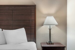 Una cama con ropa de cama blanca, un cabecero de madera, una mesita de noche con una lámpara y una pared con textura.