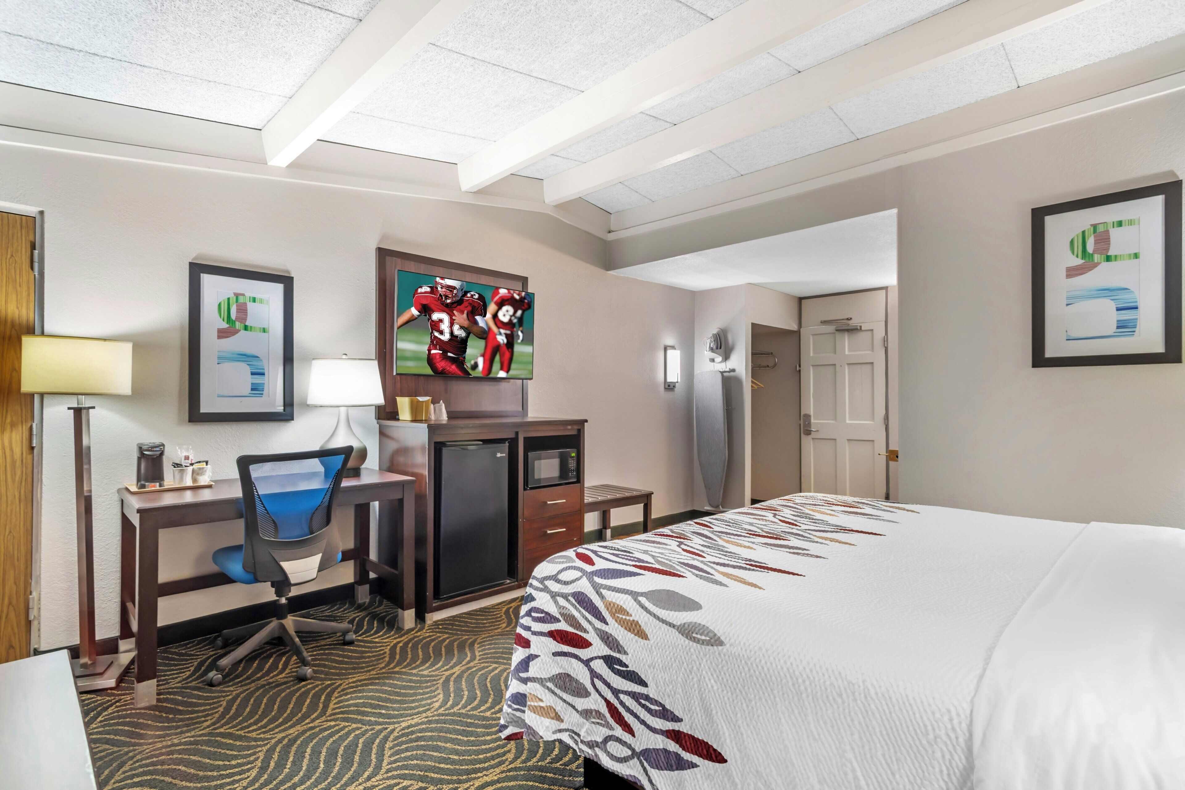 A hotel room with a bed, desk, chair, TV, and artwork on the walls.