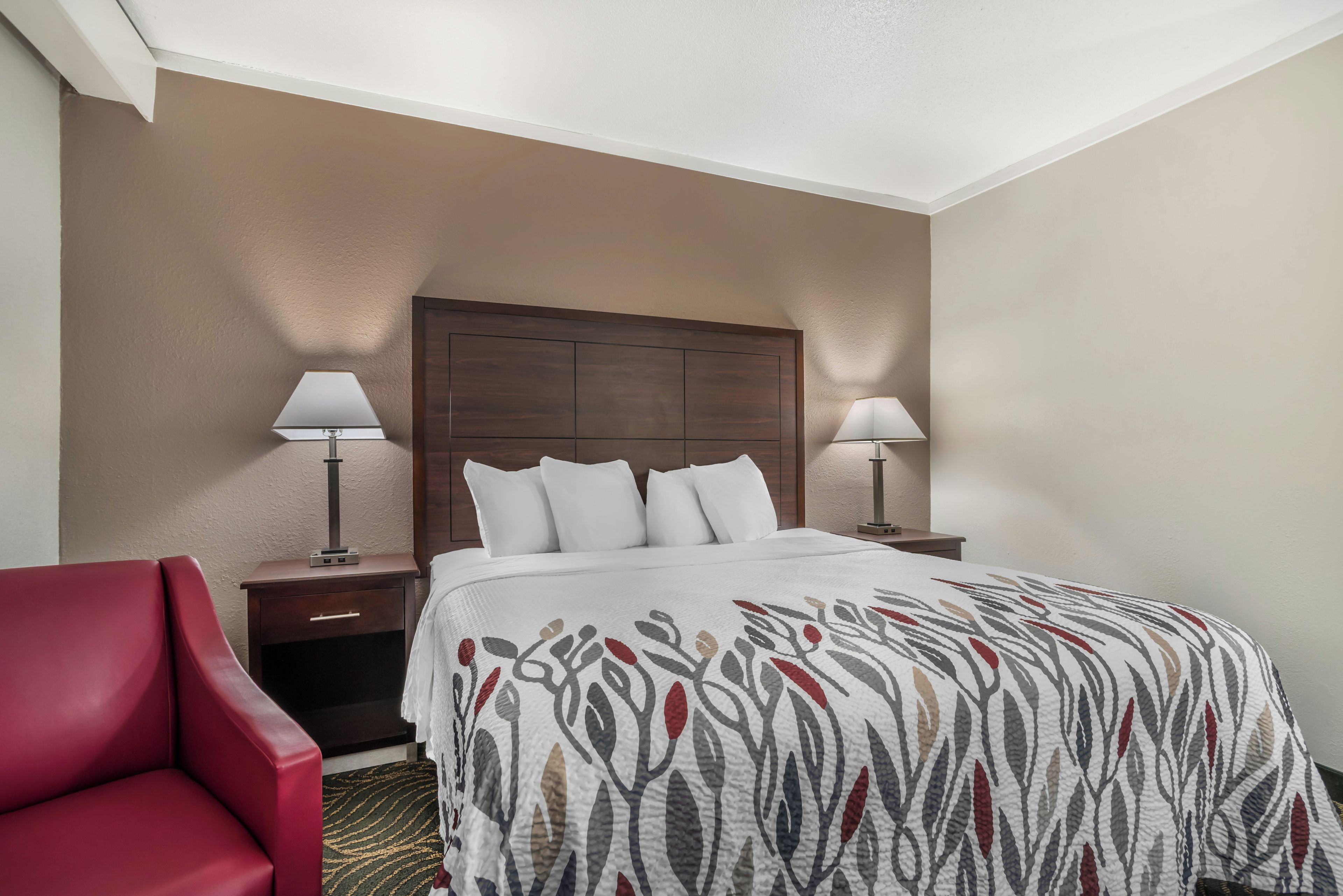 Una habitación de hotel con una cama, dos lámparas de noche, una silla roja y un cabecero de madera.