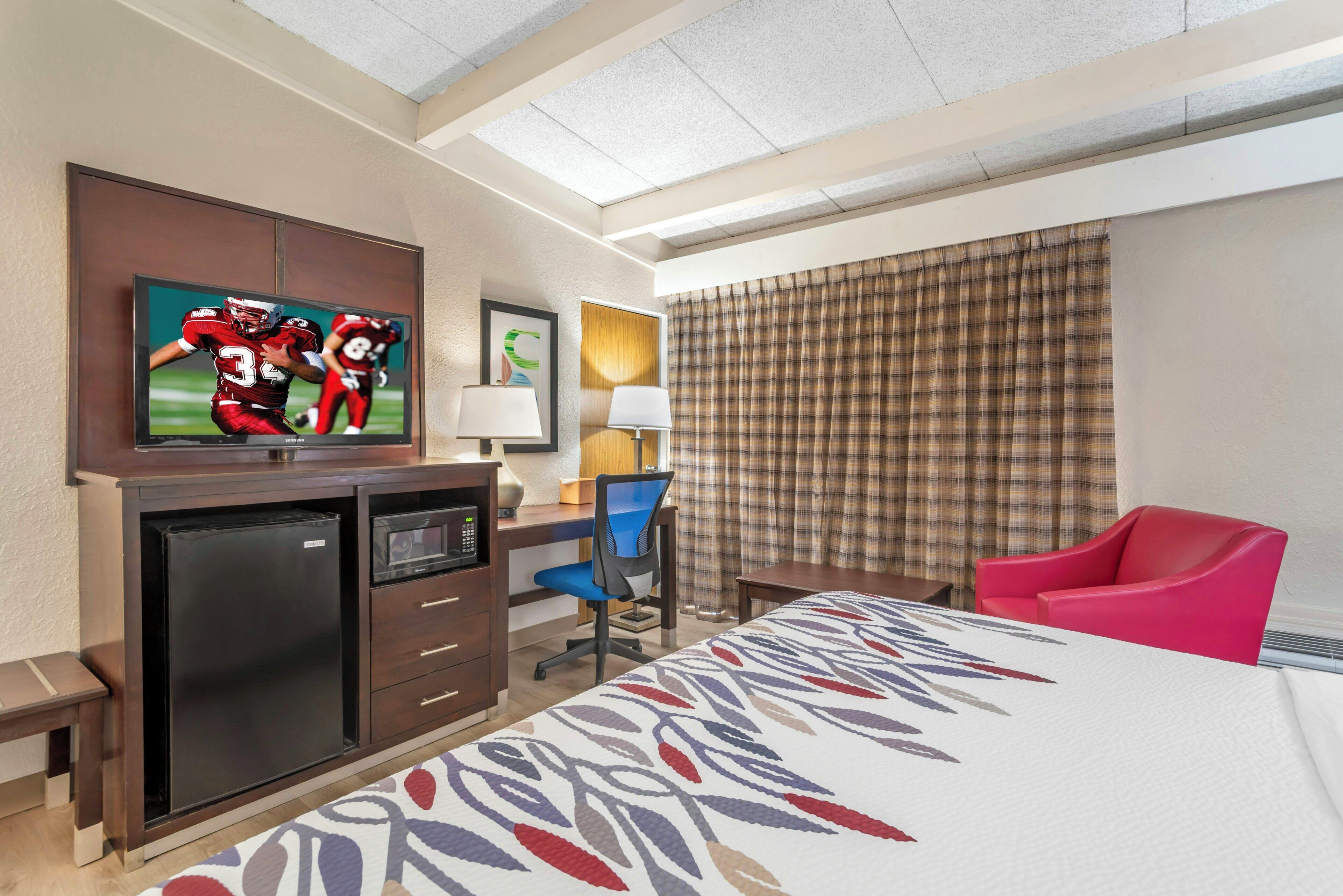 A hotel room with a bed, a desk with a computer, a television, and a red chair.