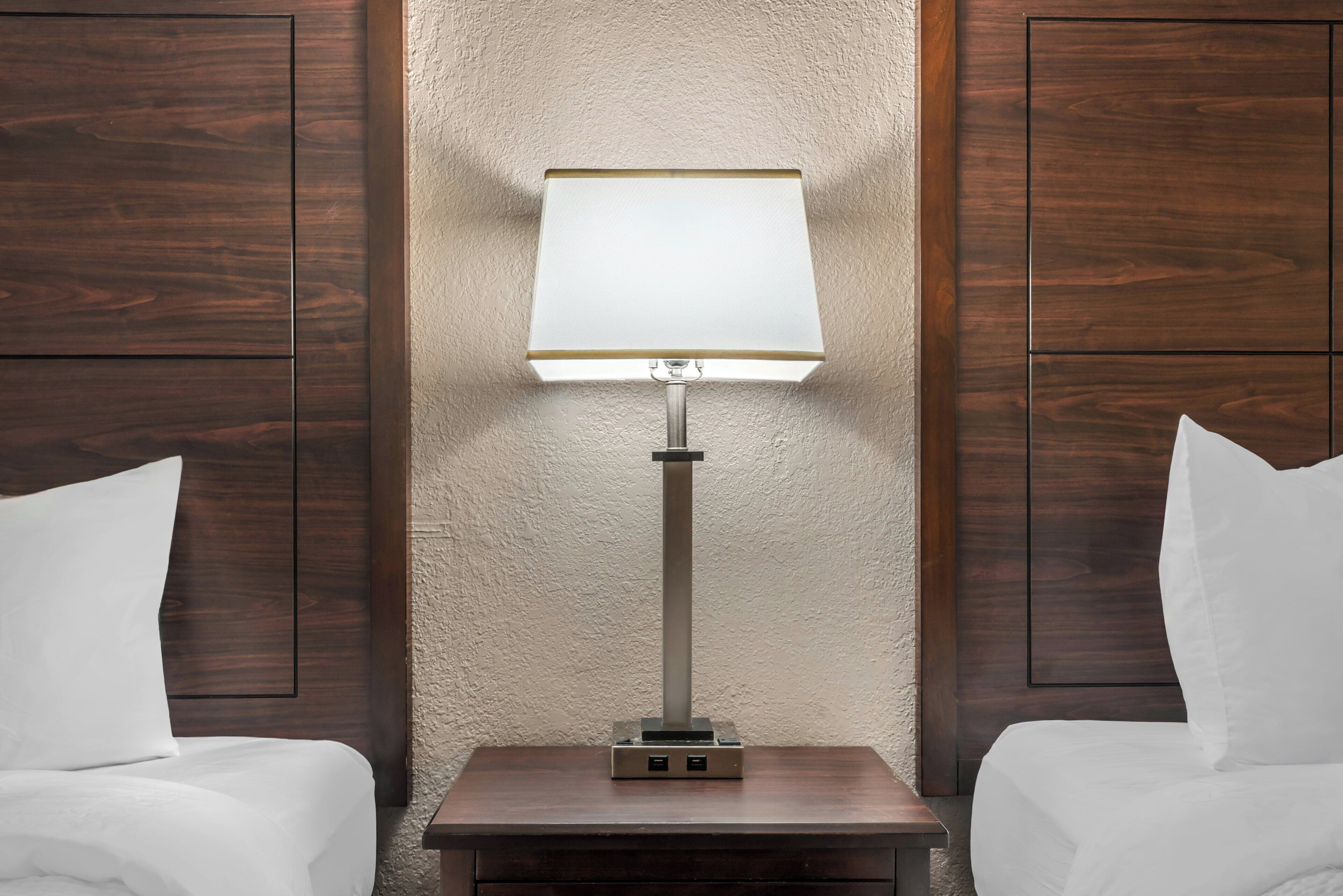 A hotel room with a bed, a nightstand with a lamp, and wooden paneling on the walls.