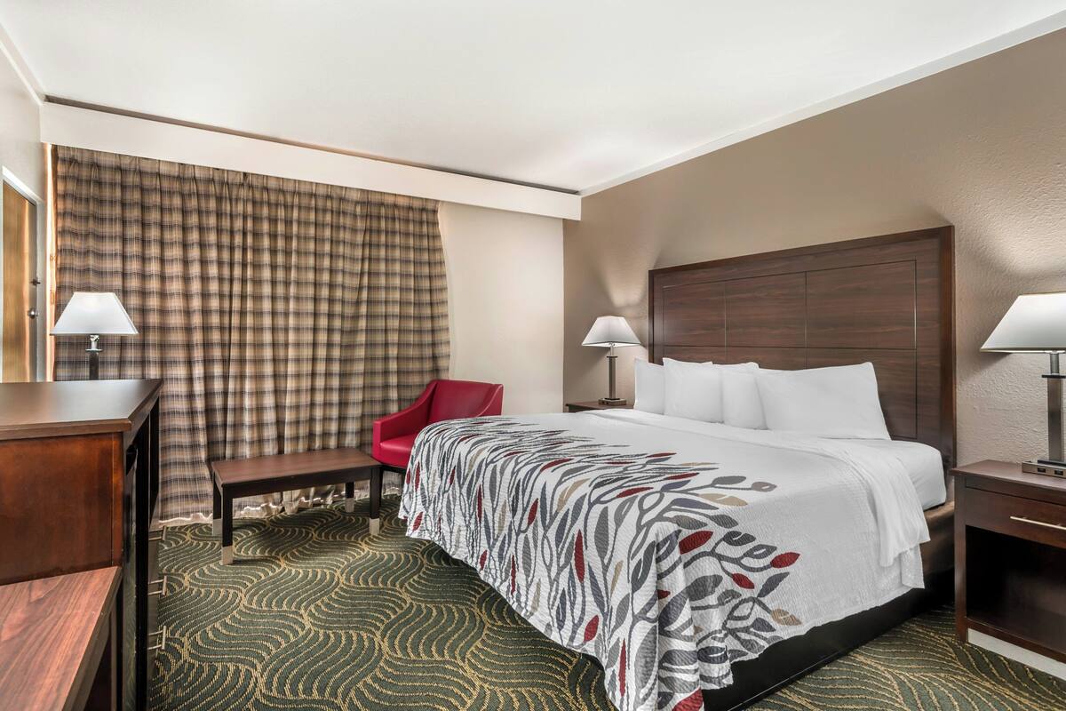 Ein Hotelzimmer mit einem Bett, zwei Nachttischen, einem Schreibtisch, einem Stuhl, einer Lampe und einem Fenster mit Vorhängen.
