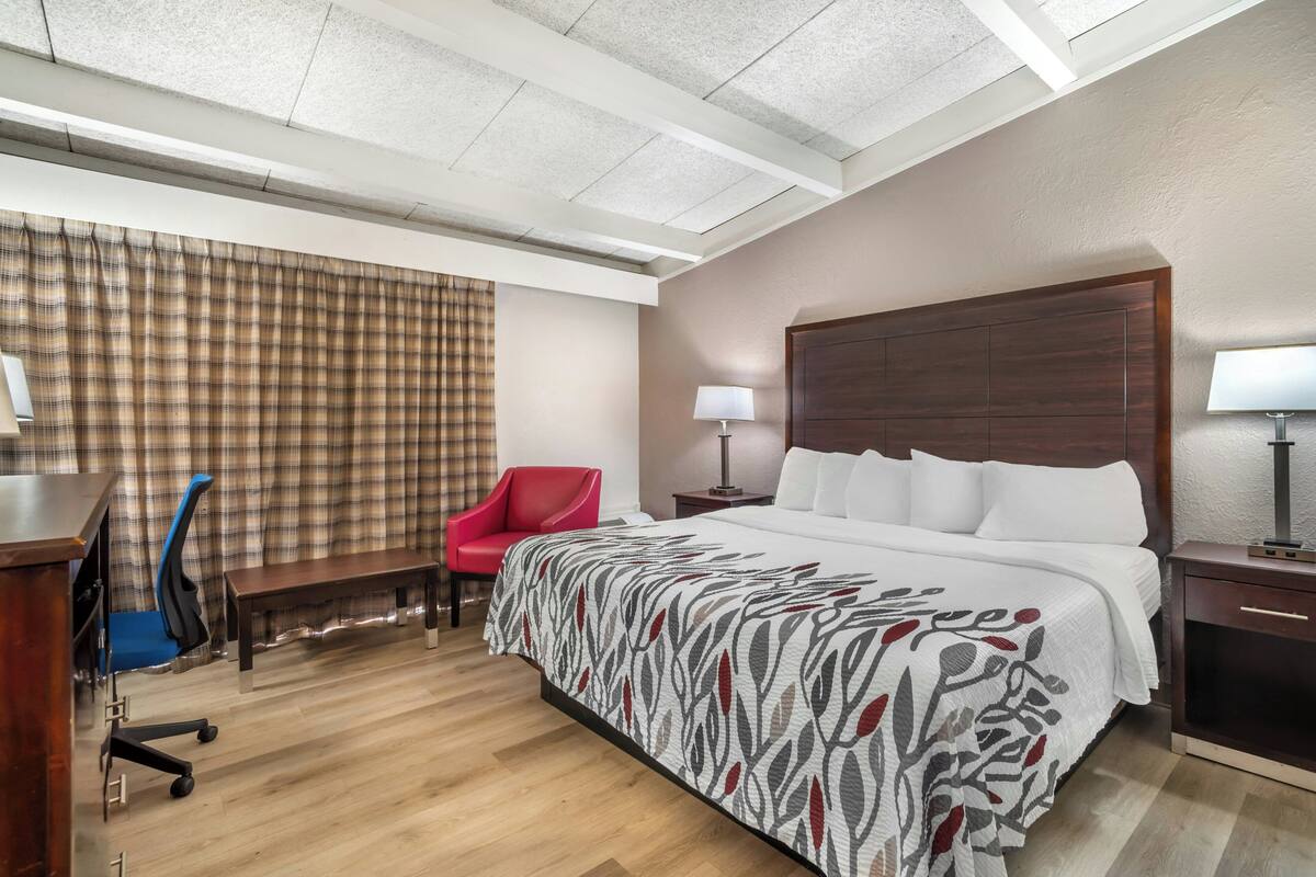 Ein Hotelzimmer mit Bett, Schreibtisch, Stuhl, Lampe und einem fensterbepflanzten Fenster.