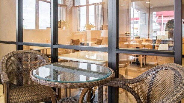A room with a glass table and wicker chairs, large windows, and a view of another room with tables and chairs.