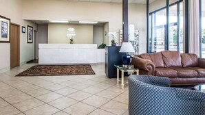 A lobby with a reception desk, a brown leather sofa, a patterned armchair, a small table with a lamp, and a large window with curtains.