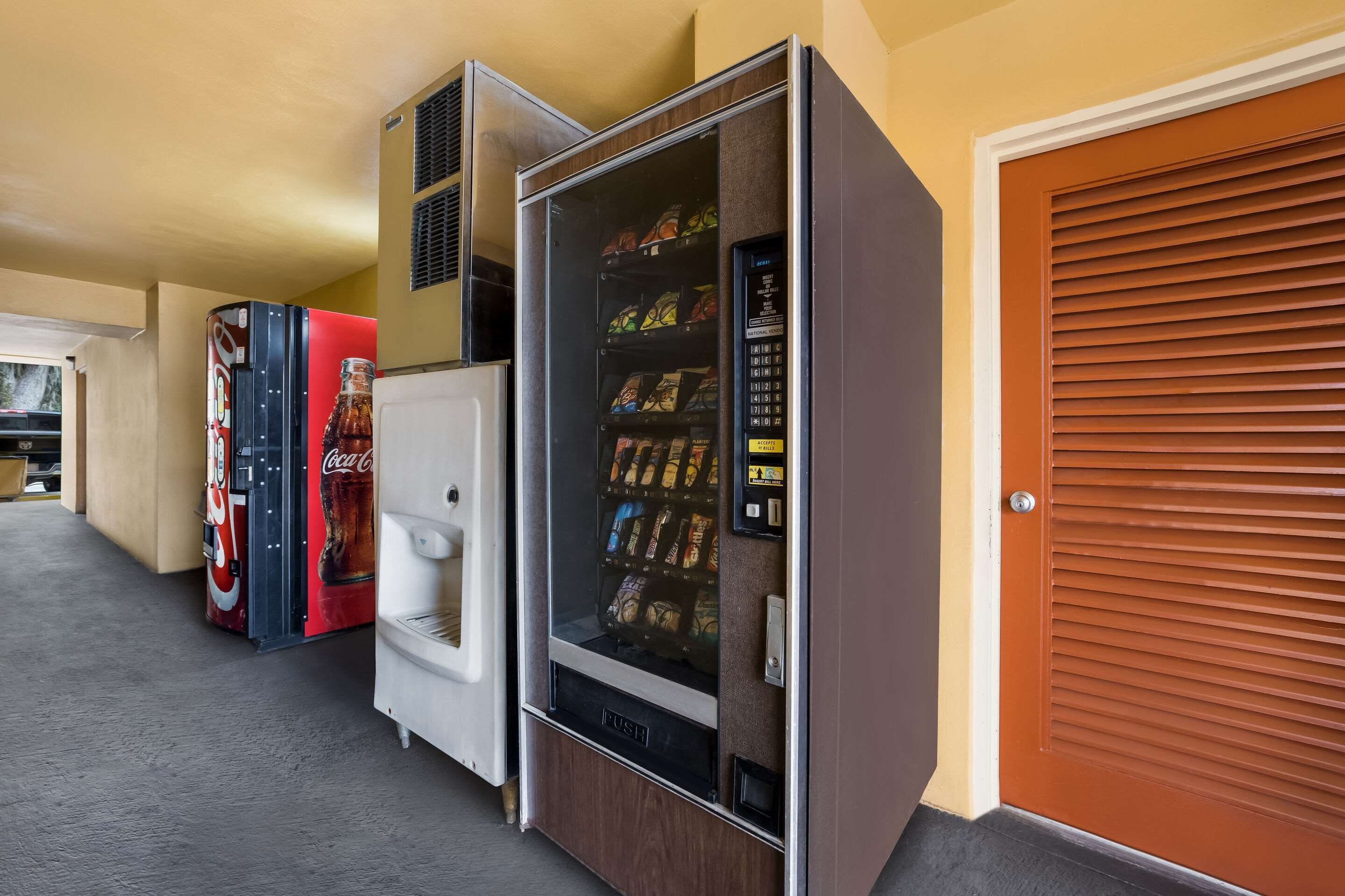 Een rij automaten met snacks en dranken, een witte koelkast en een gesloten houten deur.