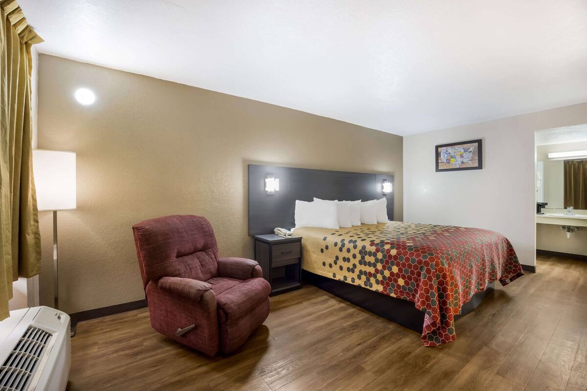 A hotel room with a bed, a red armchair, a nightstand, a lamp, and a wall-mounted picture.