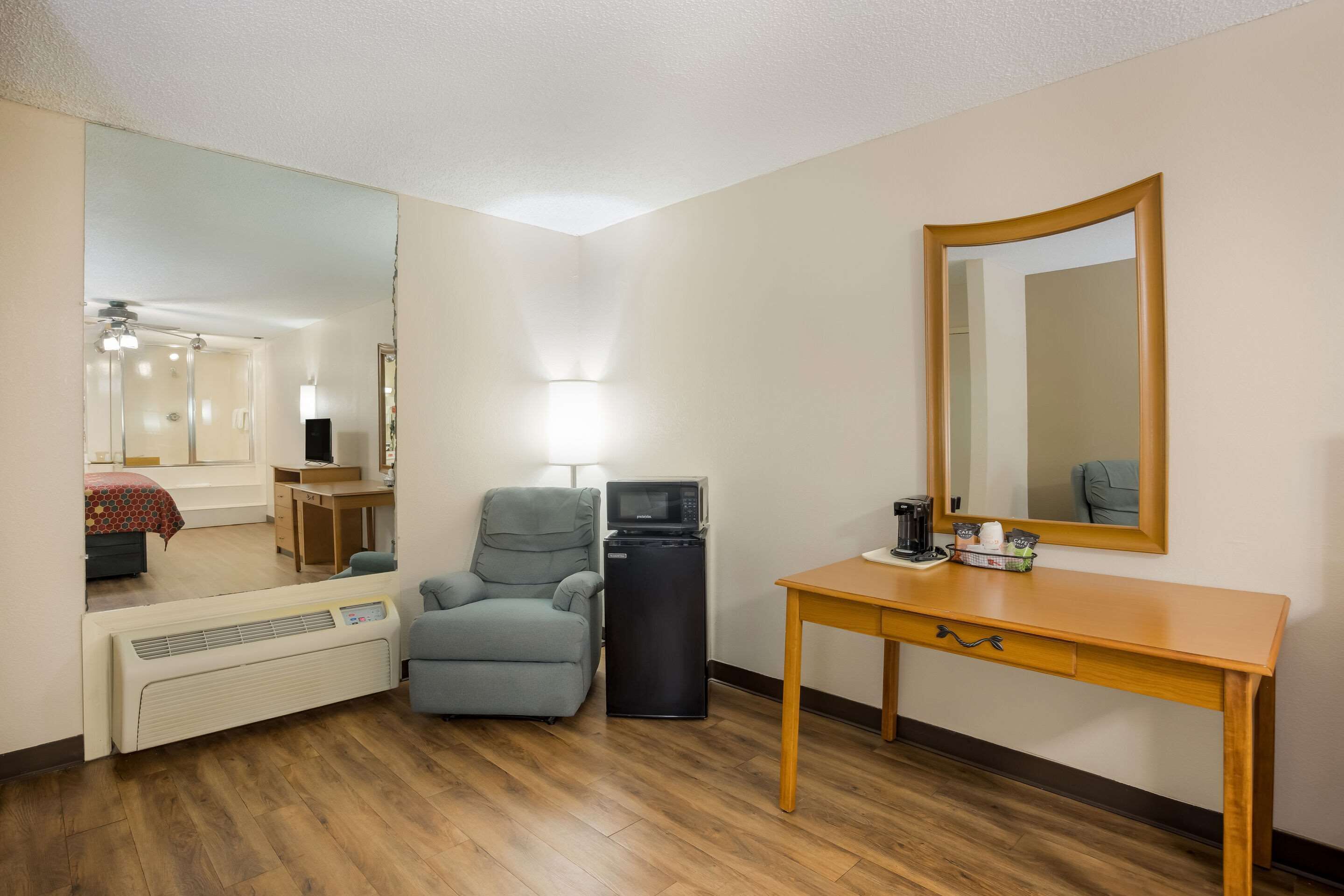 A compact hotel room with a wooden floor, a desk with a chair, a small refrigerator, a microwave, a heater, a large mirror, and a bed with a red and white patterned bedspread.