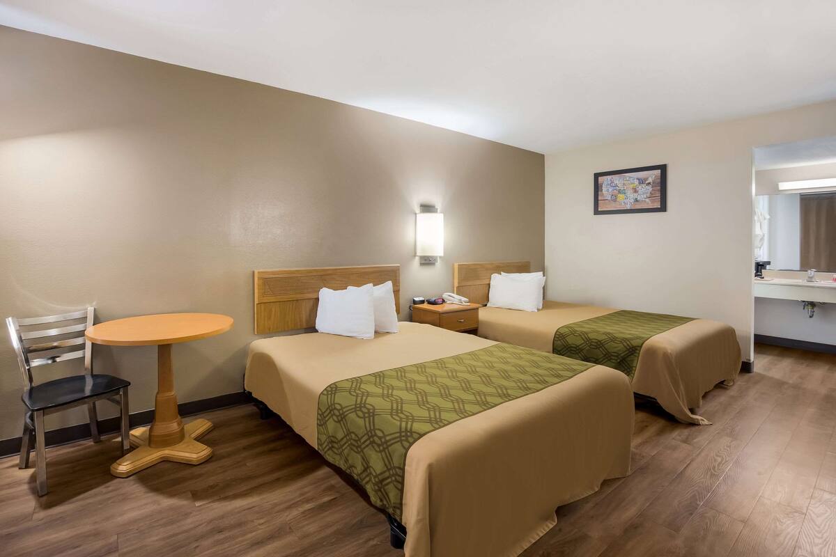 A hotel room with two beds, a small round table, a chair, a nightstand, and a wall-mounted lamp.