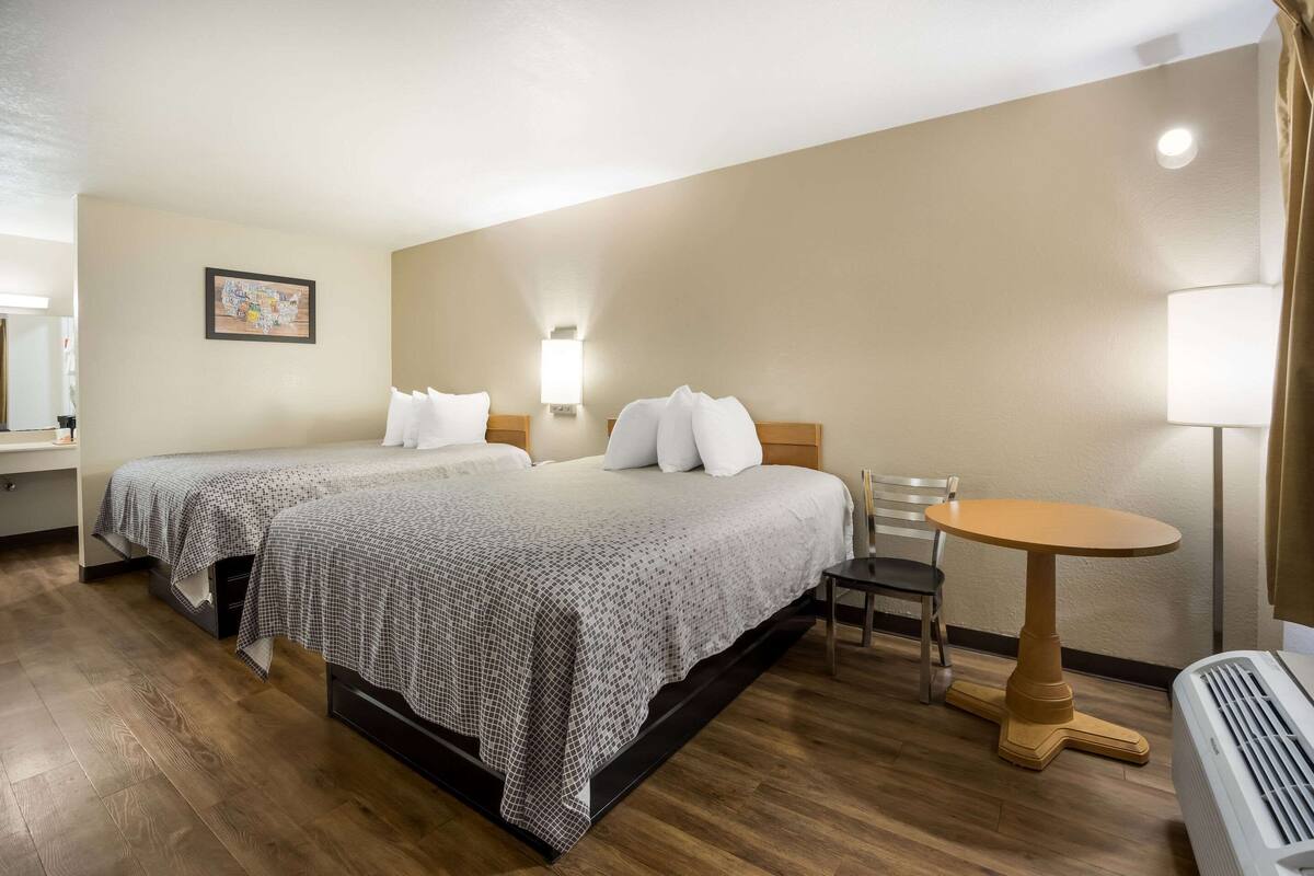 A hotel room with two beds, a small round table, a chair, a lamp, and a heater.