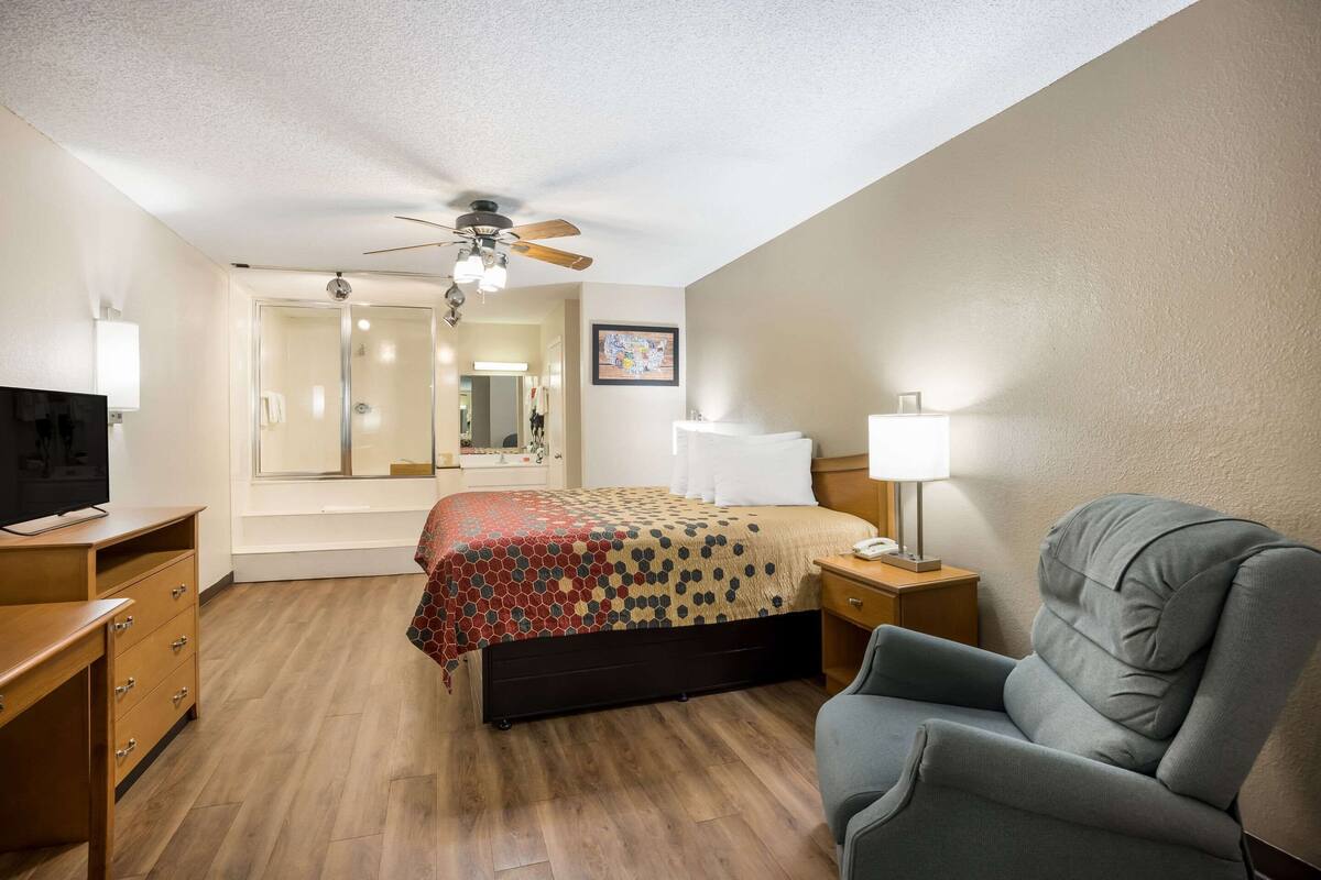 A hotel room with a bed, a desk, a chair, a TV, a ceiling fan, a mirror, and a window.