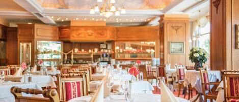 A restaurant with round tables, wooden chairs, and chandeliers.
