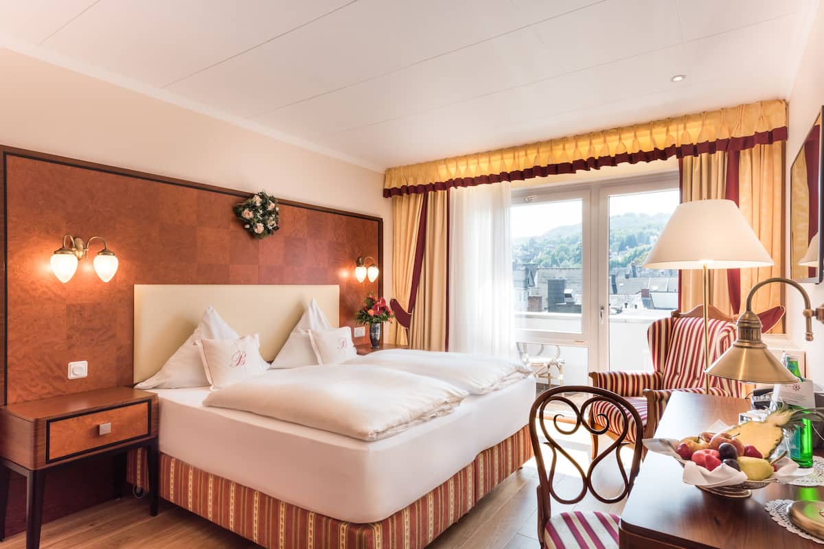 A hotel room with a large bed, two bedside lamps, a nightstand, a desk with a lamp, a chair, and a window with curtains.