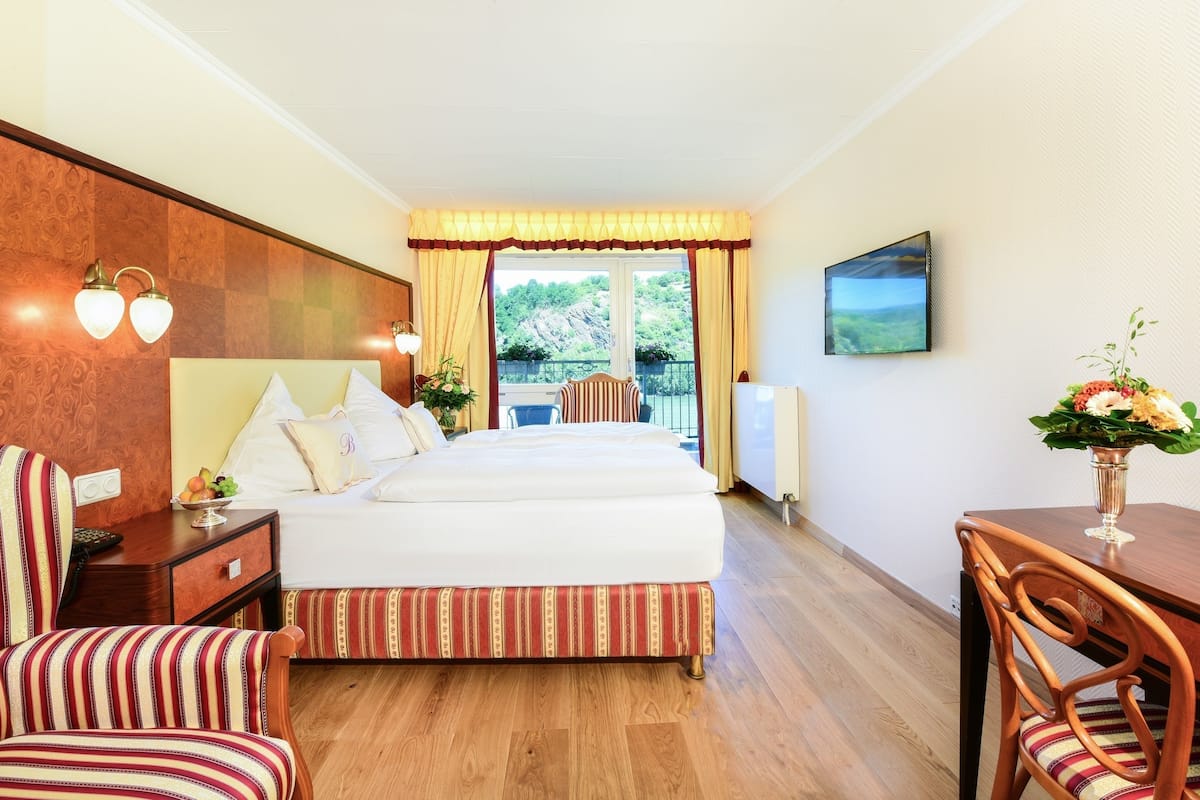 A hotel room with a bed, two bedside tables, a desk, a chair, a view of the outdoors, and a painting on the wall.