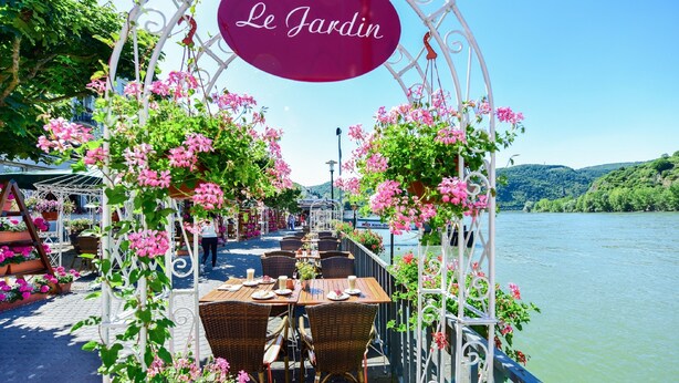 Et udendørs spiseområde med borde og stole, omgivet af lyserøde blomster og et skilt med teksten "Le Jardin".