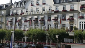 Et hotel kaldet "BELLEVUE" med flere balkoner udsmykket med blomster, i klassisk europæisk arkitekturstil, og en række parkerede biler foran.