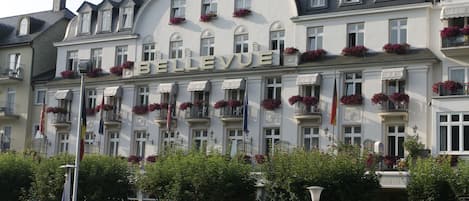A hotel named "BELLEVUE" with multiple balconies adorned with flowers, a classic European architectural style, and a row of parked cars in front.