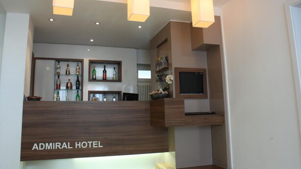 Una zona de recepción con un mostrador de madera que indica "Admiral Hotel", un televisor montado en la pared y estantes que exhiben varios objetos.