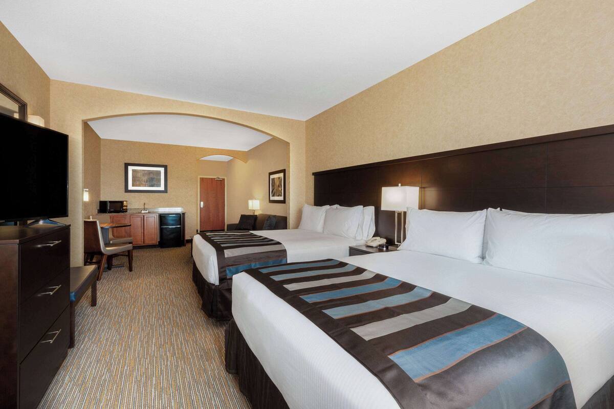 A hotel room with two beds, a desk, a TV, and a kitchenette.
