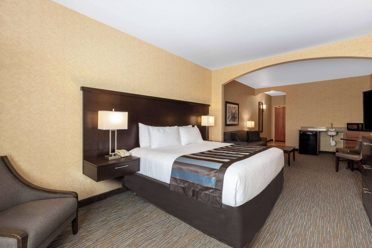 A hotel room with a large bed, bedside tables with lamps, a desk, a chair, and a kitchenette area.