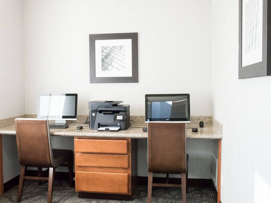 Espaço de trabalho em hotel com duas mesas, cada uma com monitor de computador, impressora e cadeira.