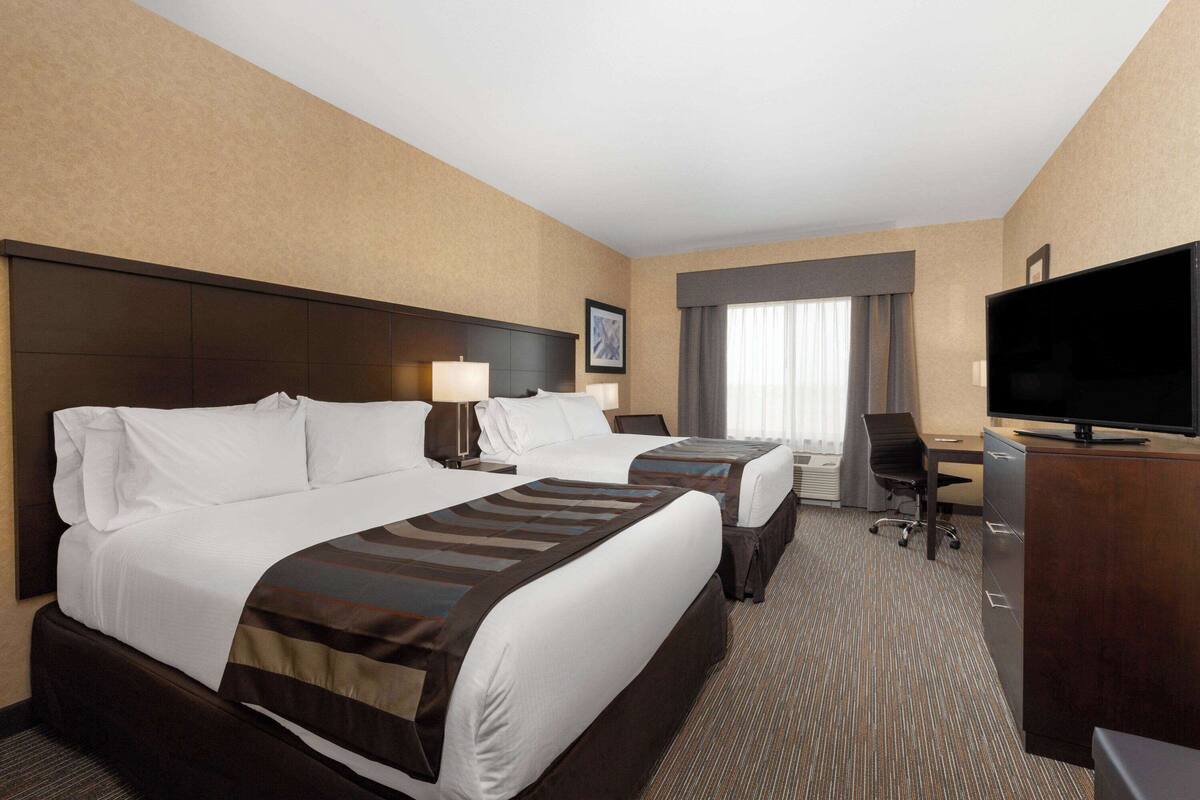A hotel room with two beds, a desk, a chair, a TV, and a large window with curtains.