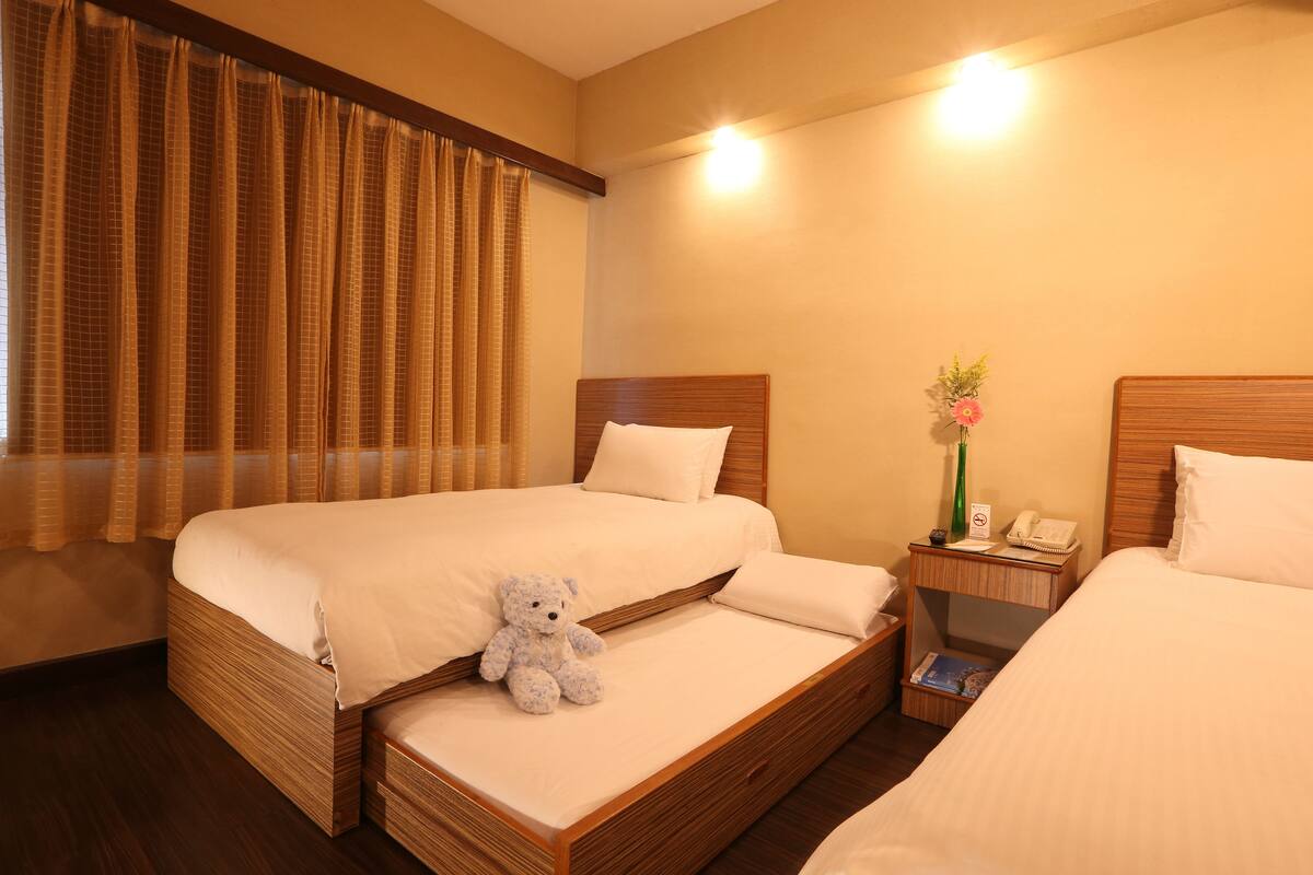 A hotel room with two beds, a nightstand with a phone and a vase of flowers, and a teddy bear on the left bed.