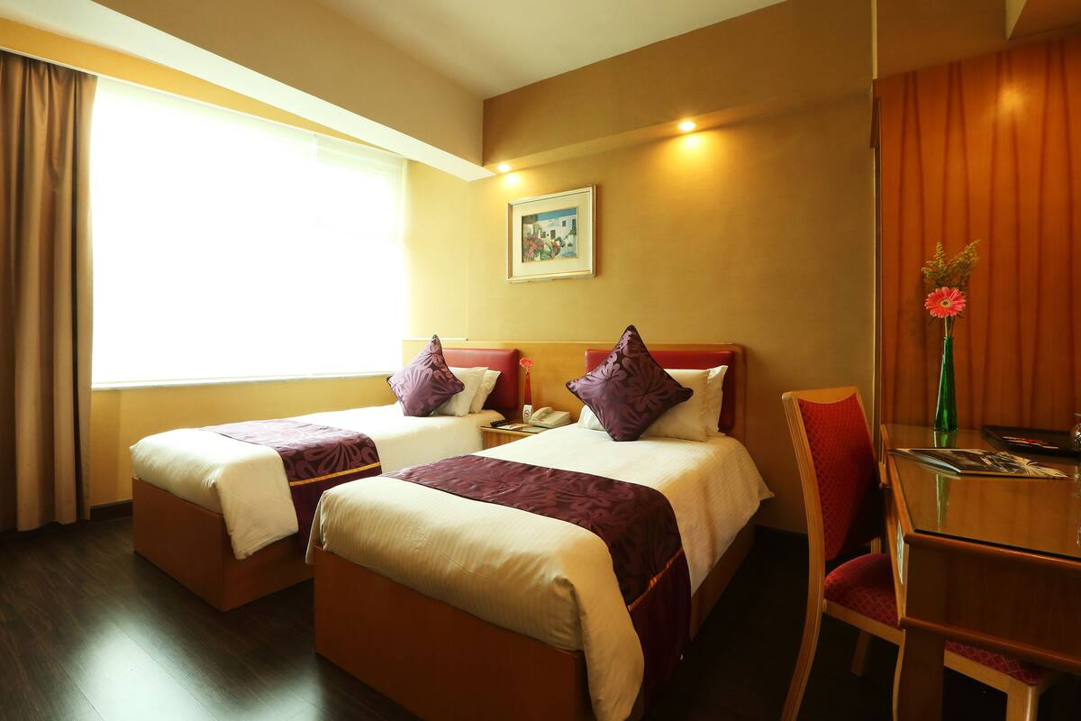 A hotel room with two beds, a desk, a chair, a window with curtains, and a painting on the wall.
