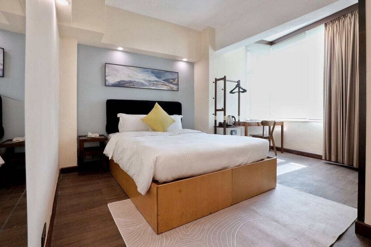 A modern hotel room with a large bed, a bedside table, a chair, a desk, and a window with curtains.