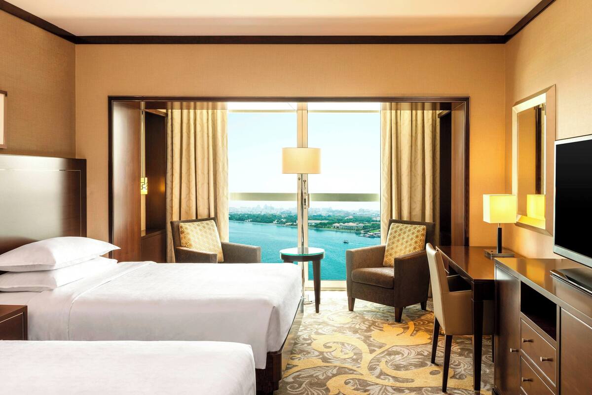 A hotel room with two beds, a desk, a TV, and a view of a body of water.