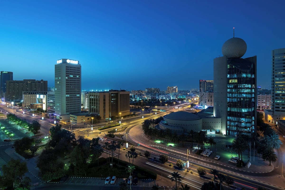 A cityscape at dusk with illuminated buildings, a roundabout, and palm trees.