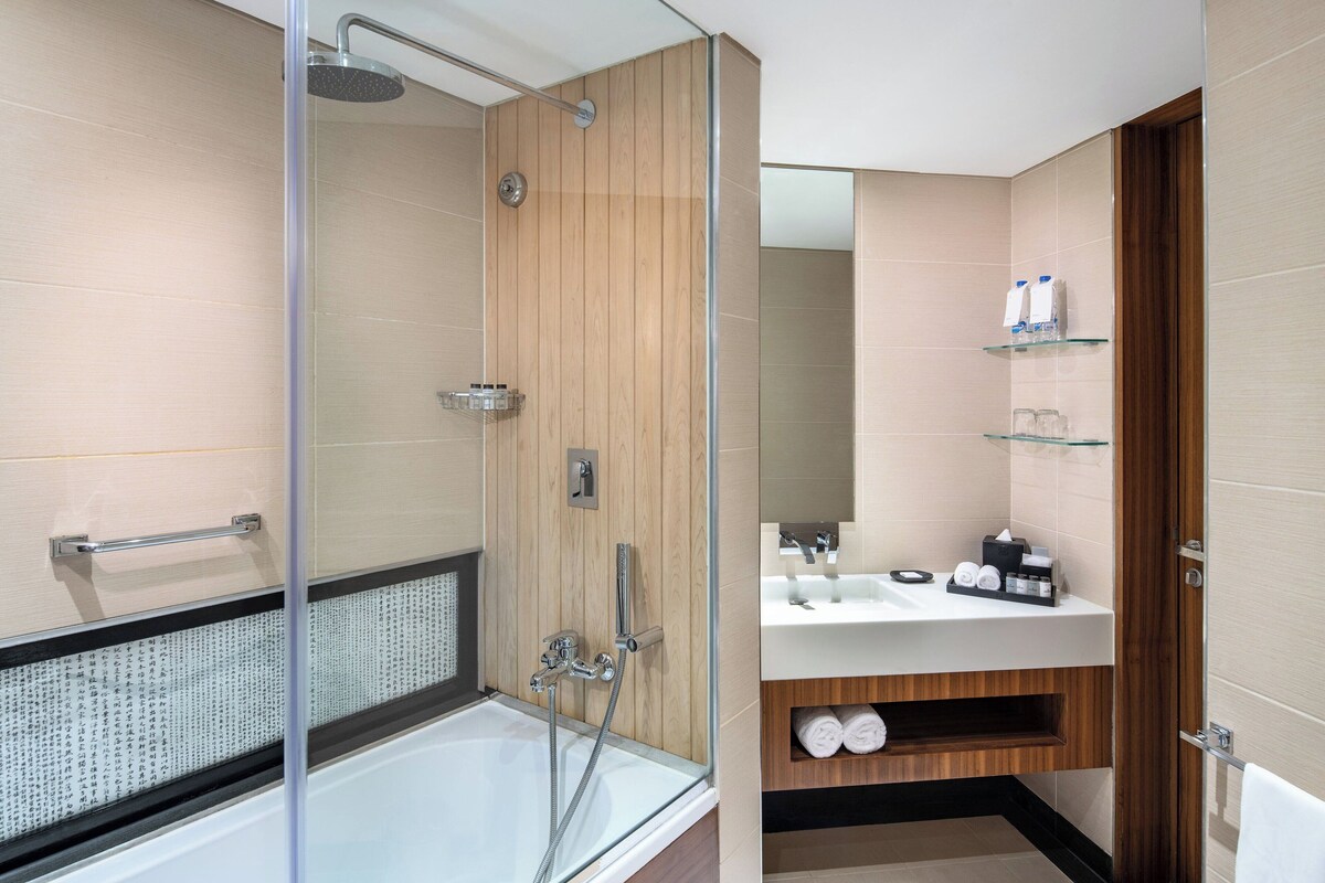 Un baño moderno con ducha de vidrio, una bañera blanca y un tocador de madera con lavamanos y perchero para toallas.