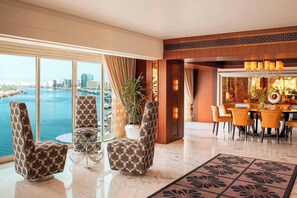 A modern hotel lobby with a large window offering a view of a body of water and cityscape. There is a seating area with patterned chairs and a glass table, and a dining area with a chandelier and orange chairs. - Sheraton Dubai Creek Hotel & Towers (Dubai)