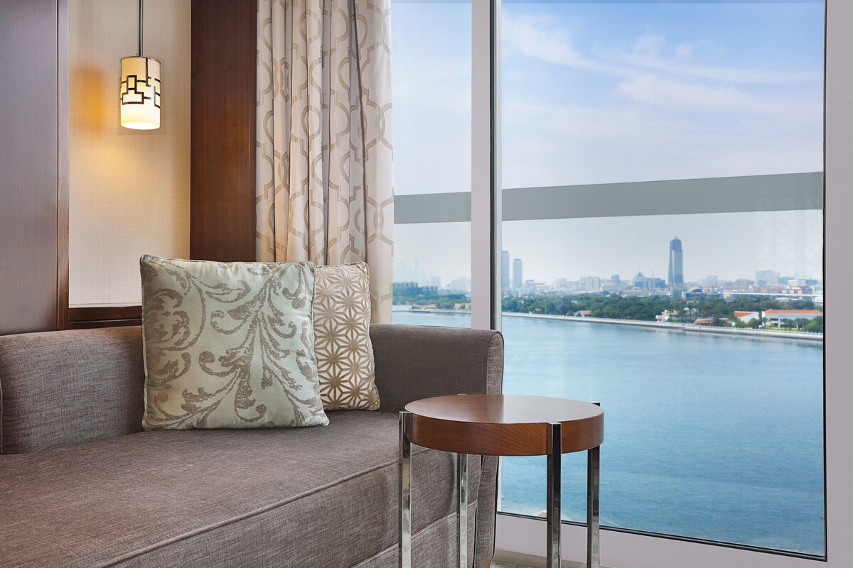Una sala de estar moderna con un sofá, una mesita redonda y vistas a un paisaje urbano y al río.