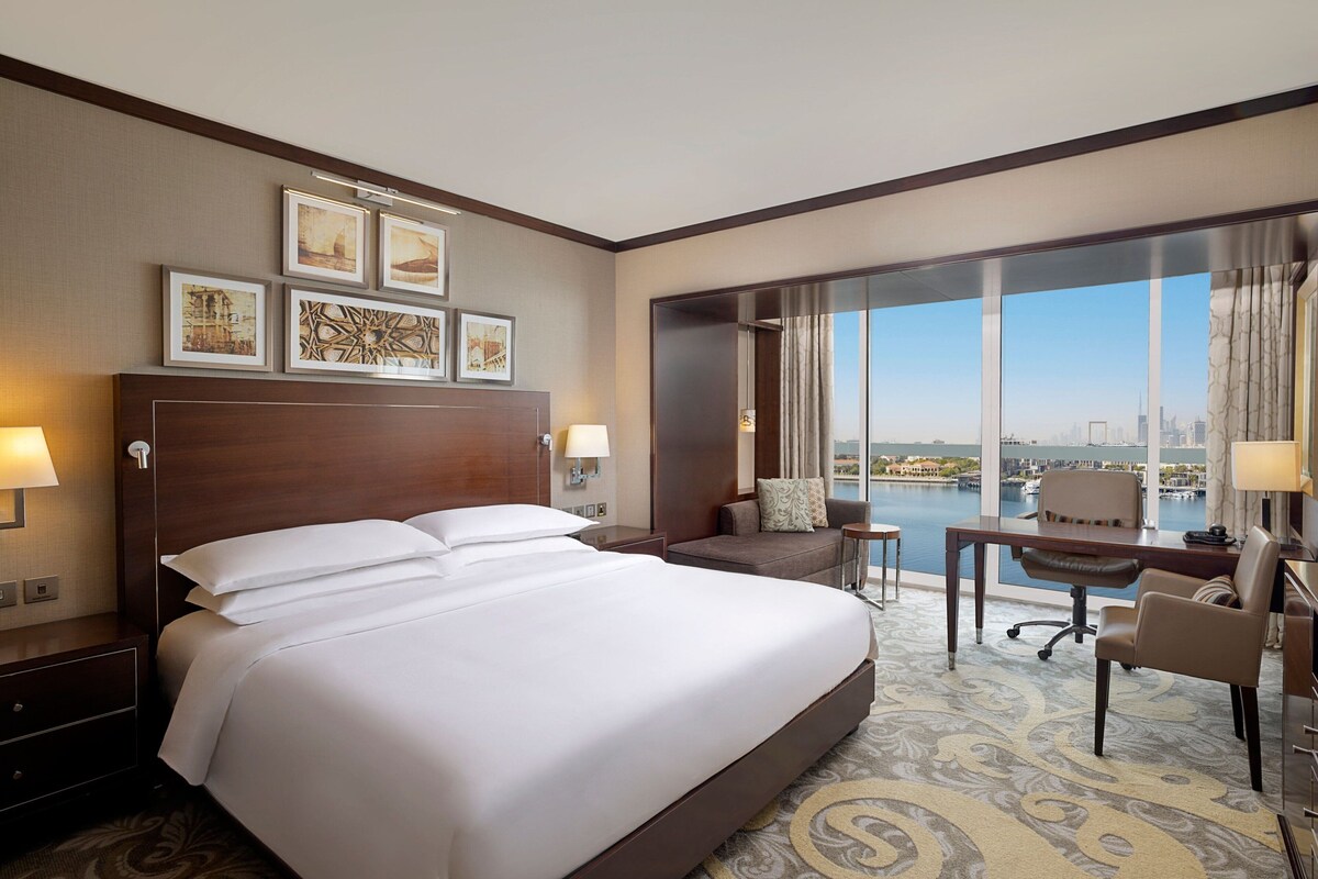 Habitación de hotel con una cama grande, un escritorio, una silla y vistas a un paisaje urbano a través de amplias ventanas.