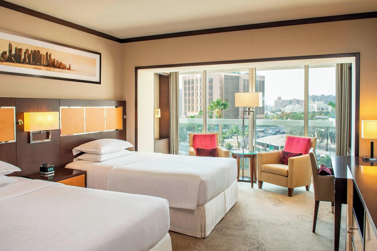 A hotel room with two beds, a desk, a chair, and a view of the cityscape through large windows.