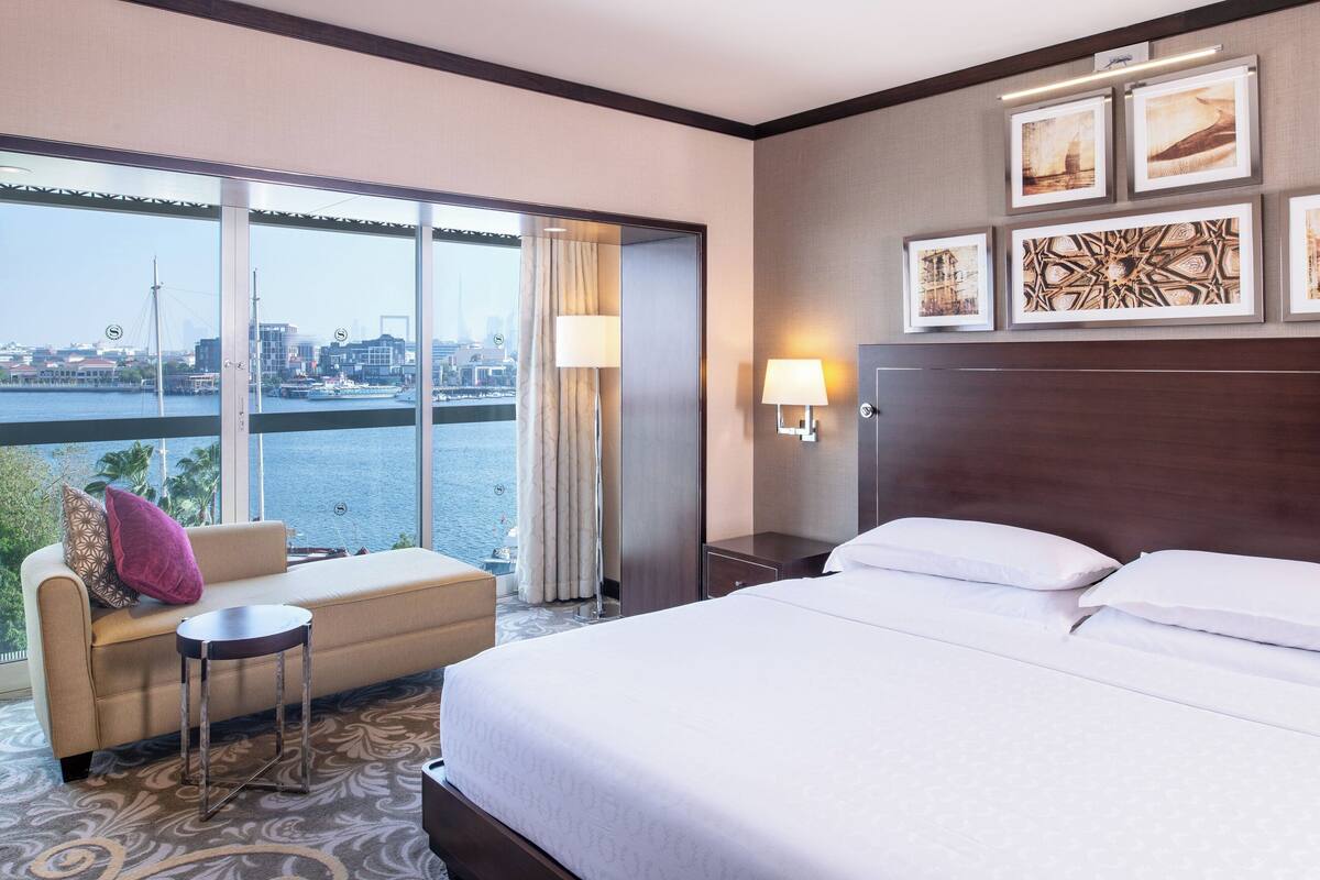 A hotel room with a bed, a sofa, a small table, and a view of a cityscape through large windows.