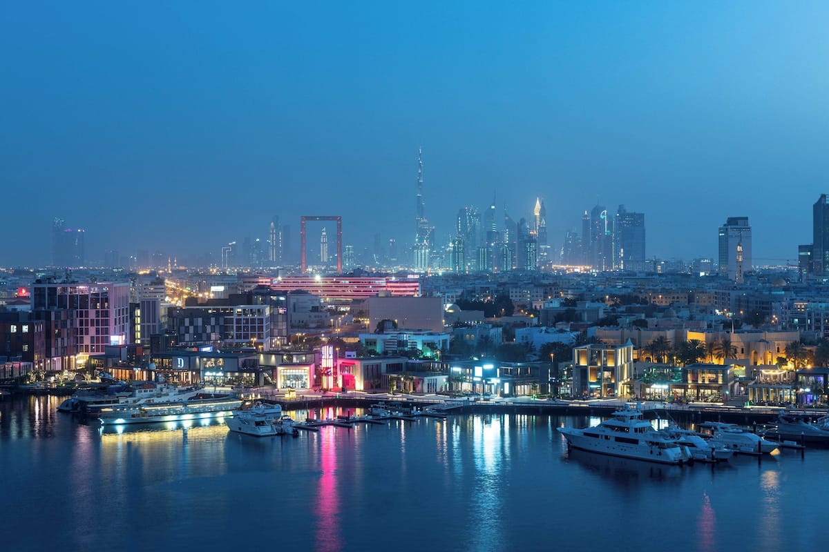 A cityscape at dusk with a marina filled with yachts, illuminated buildings, and a prominent skyscraper in the background.