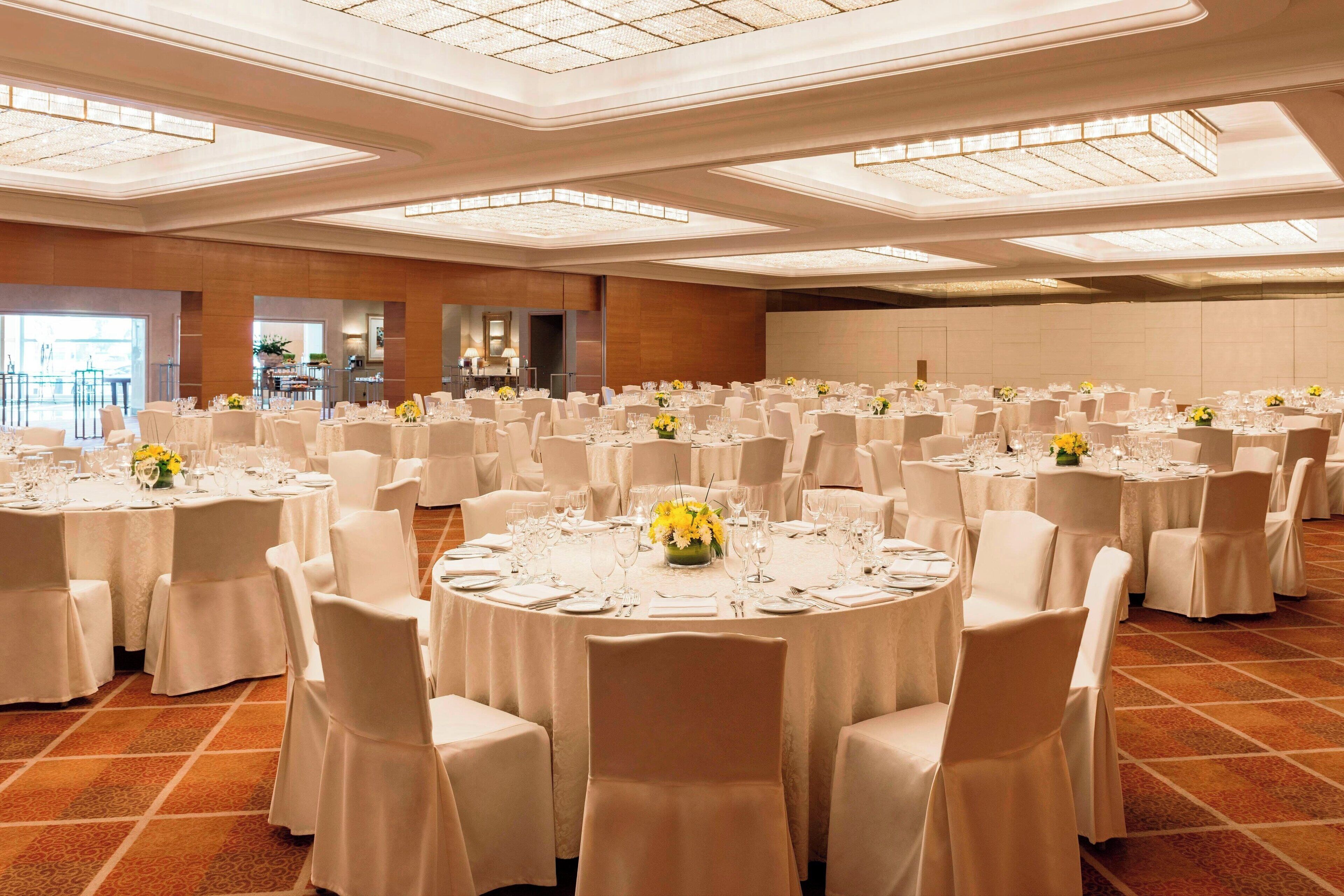 A large banquet hall with round tables set for dining, featuring white tablecloths and chairs, and a modern chandelier ceiling.