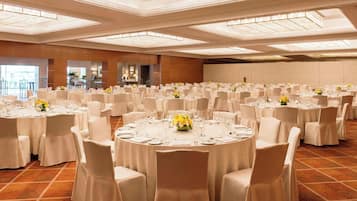 A large banquet hall with round tables set for dining, featuring white tablecloths and chairs, and a modern chandelier ceiling.