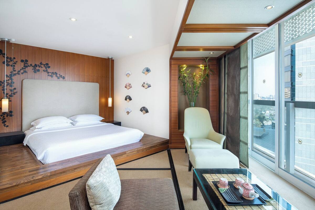 A modern hotel room with a large bed, a seating area, and a view of the city through large windows.