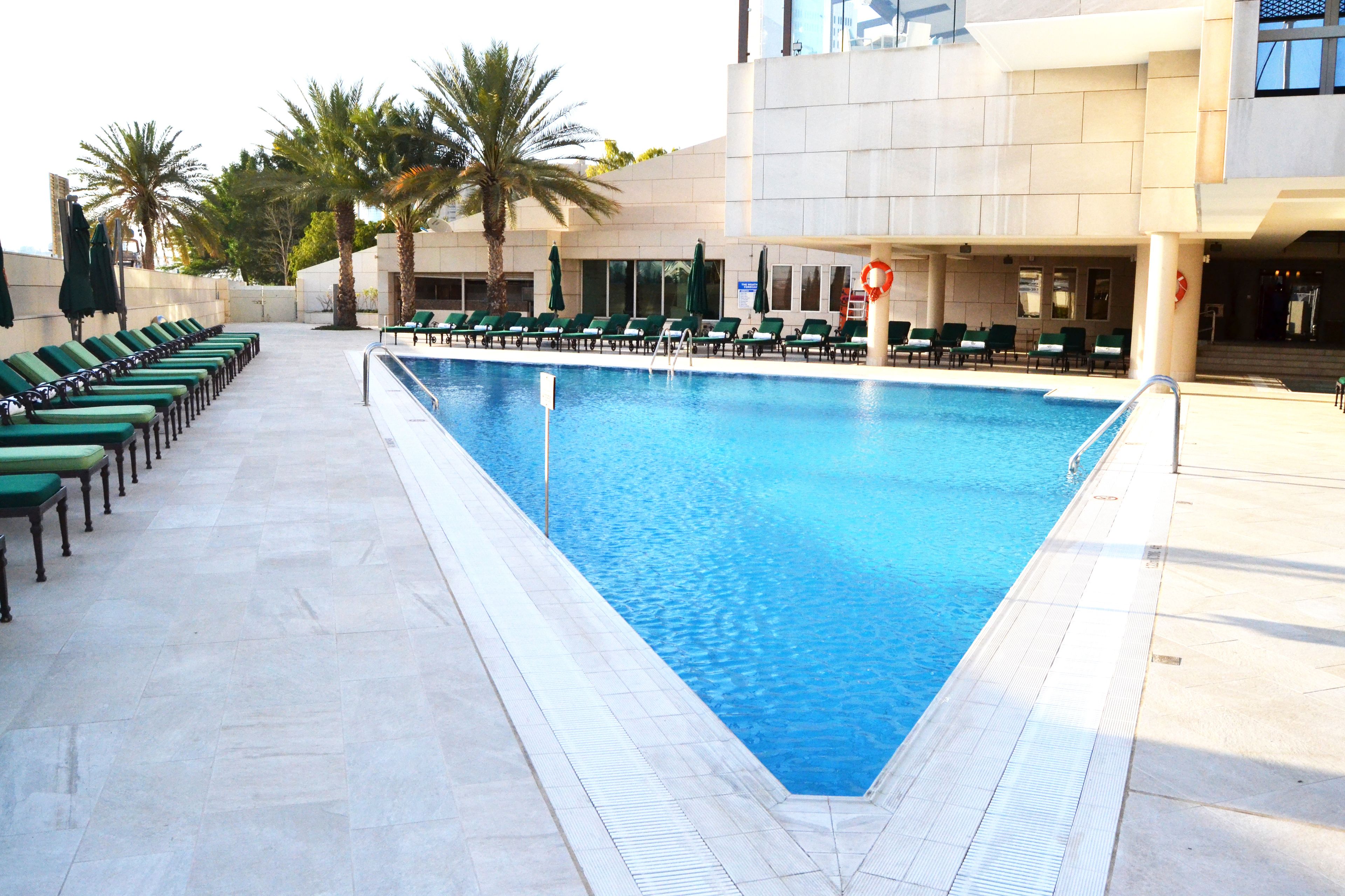A swimming pool with a tiled deck, green lounge chairs, palm trees, and a modern building in the background.