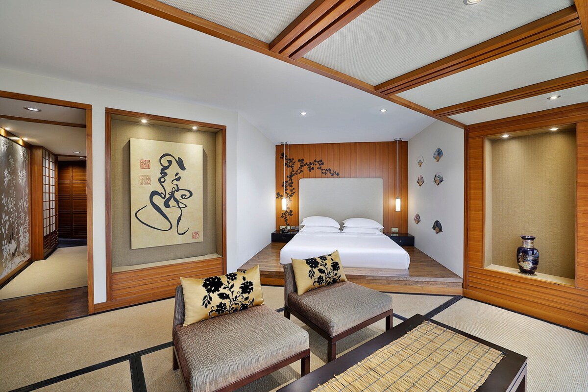 Habitación de hotel tradicional al estilo japonés con suelo de tatami, una cama grande con ropa blanca, dos bancos de tatami con cojines y una obra de caligrafía en la pared.