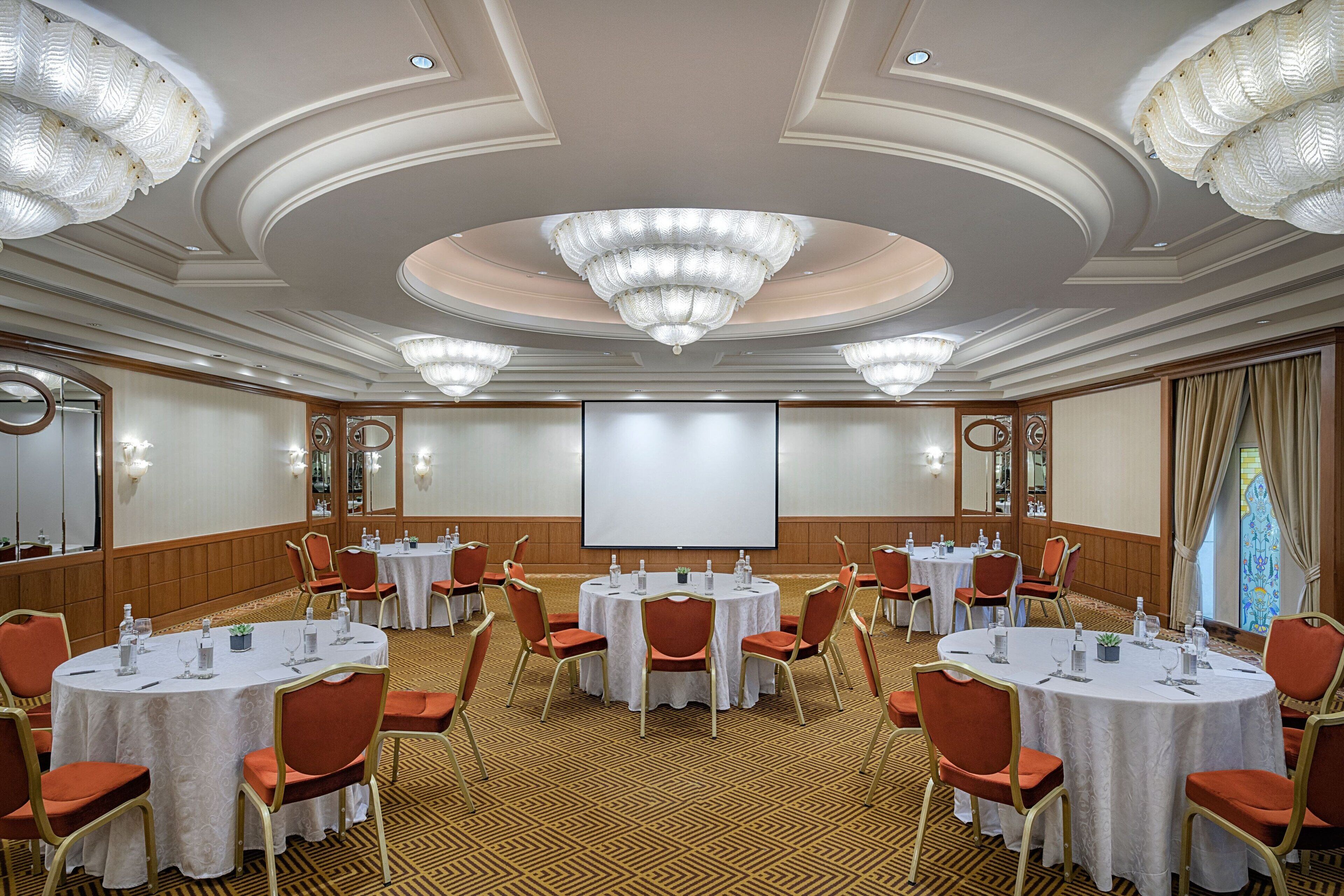 Un salón de conferencias con varias mesas redondas, sillas rojas y una pantalla grande al frente.
