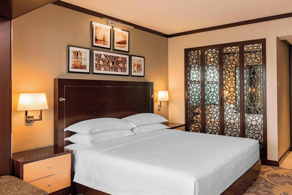 A hotel room with a large bed, two bedside lamps, a wooden headboard, a nightstand with a drawer, and a decorative screen with intricate patterns.