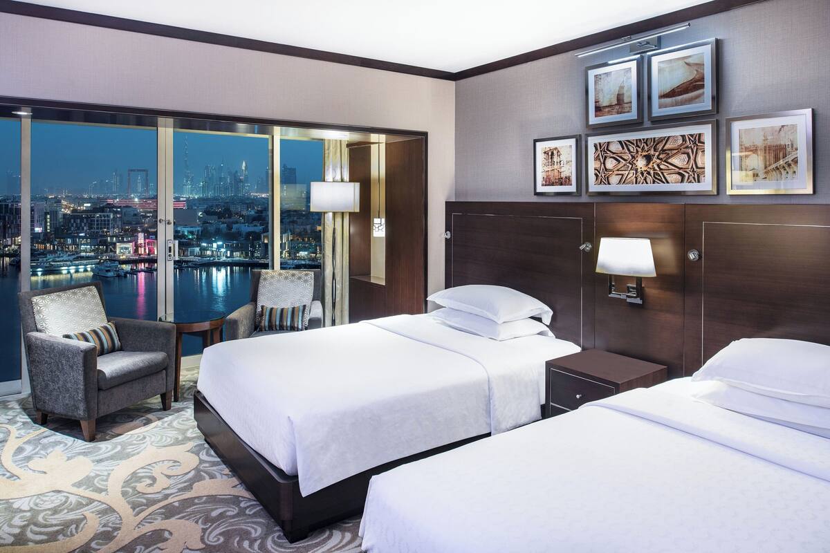 A hotel room with two beds, a chair, a nightstand, and a large window offering a city view.