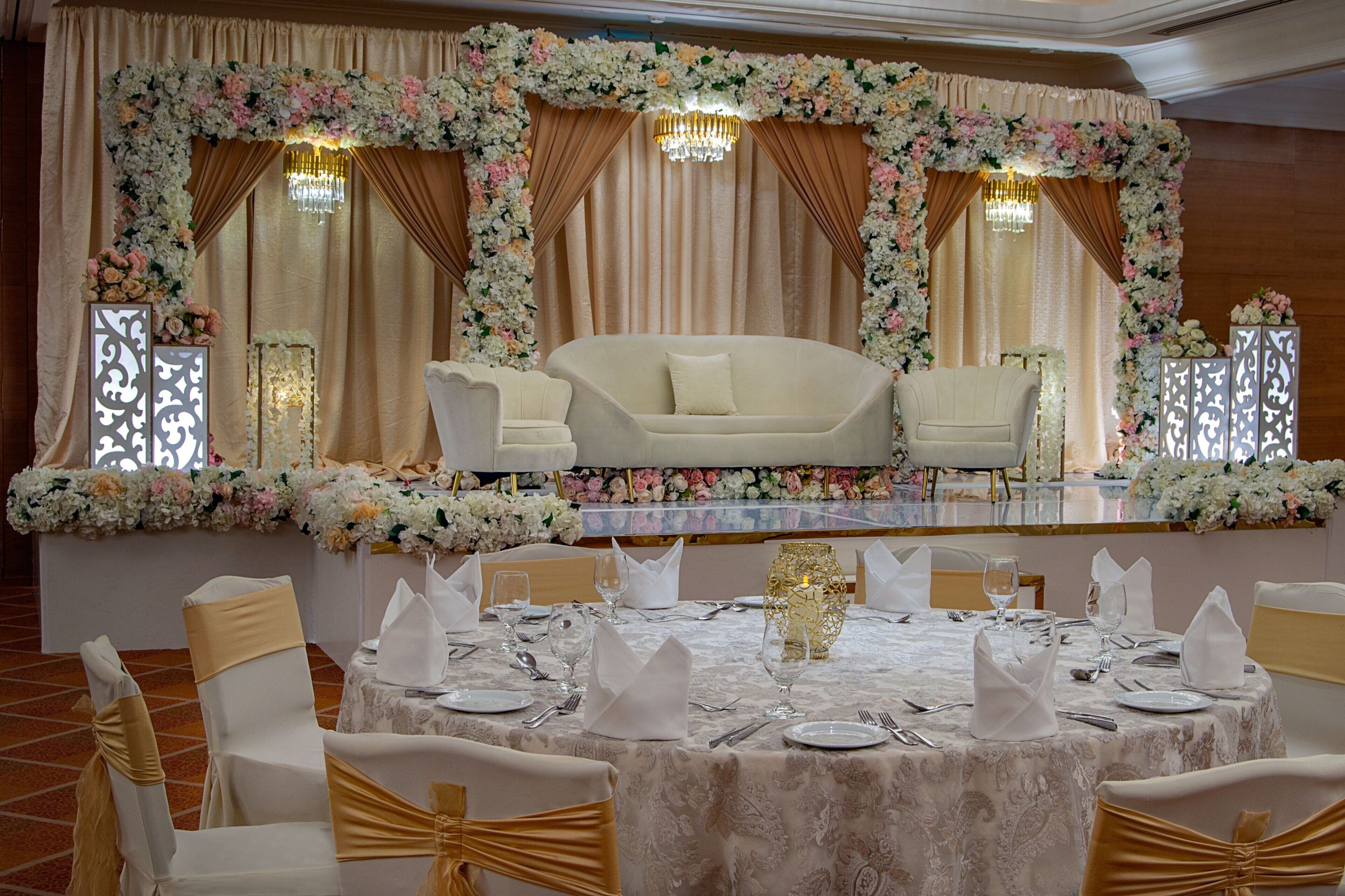 A banquet hall with a round table set for a formal event, surrounded by white chairs with gold sashes, and a backdrop of floral arrangements and drapes.