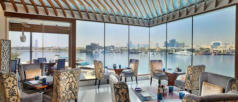 Un comedor amplio con grandes ventanales que dan a un paisaje urbano frente al agua.