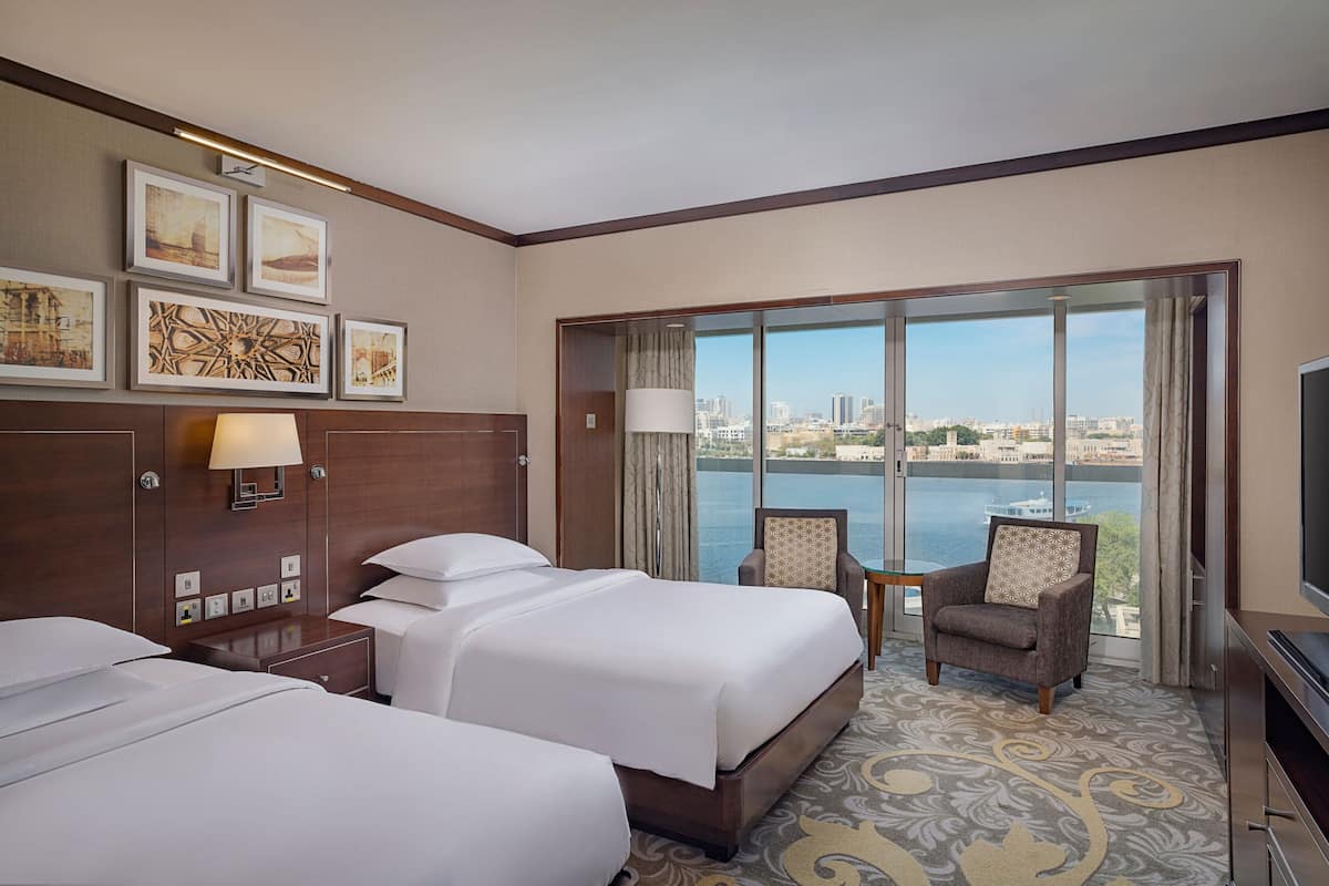 A hotel room with two beds, a TV, a chair, and a large window with a city view.
